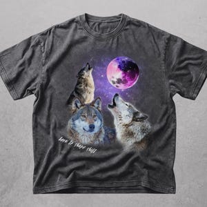 Könnte beinhalten: Ein anthrazitfarbenes Kurzarm-T-Shirt mit einem Grafikdruck von drei Wölfen und einem violetten Mond vor einem Sternenhimmel. Der Text "born to shape shift" ist in weißer Schreibschrift unter den Wölfen auf der Vorderseite aufgedruckt.