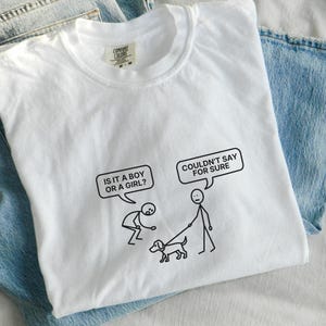 Peut inclure: T-shirt blanc avec un dessin humoristique de personnages bâtons. Le graphisme comprend des bulles de dialogue avec les phrases "IS IT A BOY OR A GIRL?" et "COULDN'T SAY FOR SURE". Le t-shirt est plié sur un jean bleu.