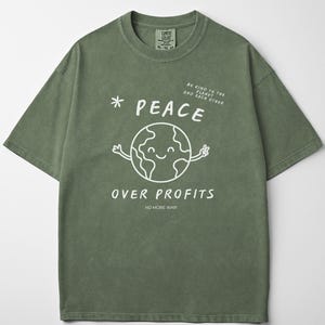 May include: An olive green t-shirt featuring a white graphic of a smiling Earth with outstretched arms, the word "PEACE" above it, and "OVER PROFITS" below. The text "BE KIND TO THE PLANET AND EACH OTHER" and "NO MORE WAR" are also printed on the shirt.