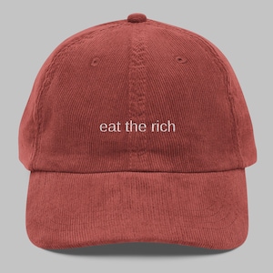 May include: A brick-red corduroy baseball cap featuring the embroidered phrase "eat the rich" in white. The cap has a curved peak and a rounded crown, suitable for casual wear.