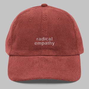 Op de afbeelding: Roodbruine corduroy baseballcap met de woorden "radical empathy" in het wit geborduurd. De cap heeft een gebogen klep en een ronde kroon, geschikt voor casual wear.