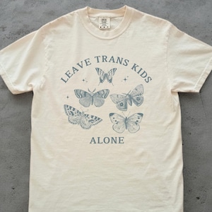 Puede incluir: Una camiseta de color crema con el texto "LEAVE TRANS KIDS ALONE" impreso en azul. El texto está rodeado de cinco ilustraciones de mariposas.