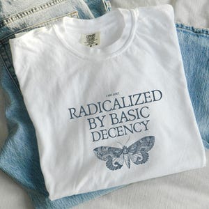 Könnte beinhalten: Weißes Comfort Colors T-Shirt mit dem Text "I AM JUST RADICALIZED BY BASIC DECENCY" und einer dunkelblauen Mottenillustration. Das T-Shirt liegt auf einer hellblauen Jeans.