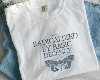 May include: White Comfort Colors t-shirt with the text "I AM JUST RADICALIZED BY BASIC DECENCY" and a moth illustration in navy blue. The shirt is laid on top of a pair of light blue denim jeans.
