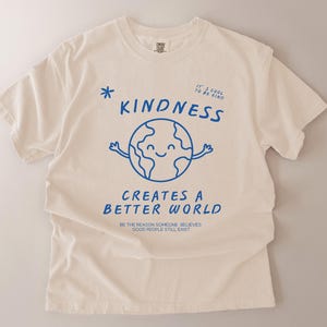 May include: Cream-coloured t-shirt with blue text and graphics. The shirt displays the word "KINDNESS" above a smiling globe with arms. Below, it reads "CREATES A BETTER WORLD" and "BE THE REASON SOMEONE BELIEVES GOOD PEOPLE STILL EXIST."