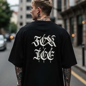 May include: Black oversized t-shirt with white gothic lettering on the back. The shirt has short sleeves and a relaxed fit. The person is walking down a city street.