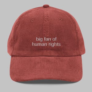 May include: A brick-red corduroy baseball cap featuring the embroidered phrase "big fan of human rights" in white. The cap has a curved peak and a rounded crown with a button. A classic six-panel design, suitable for various head sizes.