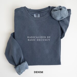 Pode incluir: Um moletom azul denim com o texto "RADICALIZED BY BASIC DECENCY" em branco. O moletom tem gola redonda e mangas compridas, com a manga de uma manga enrolada. A palavra "DENIM" é impressa na parte inferior.