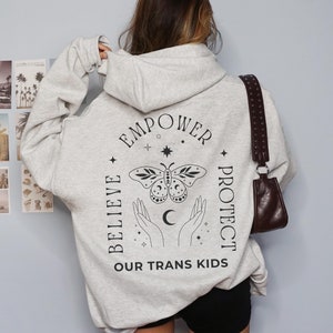 Può includere: Una felpa grigia con una grafica di farfalla nera e il testo "EMPOWER BELIEVE PROTECT OUR TRANS KIDS".