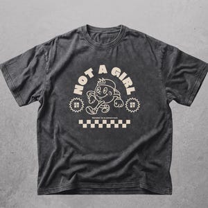 Könnte beinhalten: Dunkelgraues T-Shirt mit cremefarbenem Aufdruck. Der Aufdruck zeigt die Worte "NOT A GIRL" über einer Cartoonfigur. Darunter steht der Text "Gender is a construct" und ein cremefarbenes und weißes Schachbrettmuster.