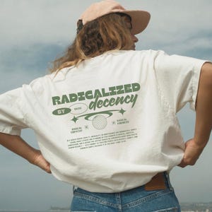 Könnte beinhalten: Ein cremefarbenes T-Shirt mit dunkelgrünem Text auf der Rückseite, der RADICALIZED decency, BY BASIC, RADICAL EMPATHY und RADICAL KINDNESS lautet, sowie einer Grafik einer Discokugel, Sternen und einem Absatz mit kleinem Text.
