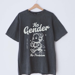 Può includere: Una maglietta grigia scura con una grafica bianca di un orso dei cartoni animati che indossa delle cuffie e tiene una bevanda. Il testo "No Gender" è scritto in un grande font stilizzato sopra l'orso. Il testo "Just Be You" è scritto verticalmente sul lato sinistro dell'orso. Il testo "No Problem" è scritto sotto l'orso.