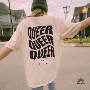 May include: Cream-colored t-shirt with the word "QUEER" stacked three times in a wavy, black font. Below the text is the word "CLUB" and two small star designs. The shirt is part of the Kind Find Apparel collection.