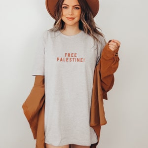 K&ouml;nnte beinhalten: Ein graues T-Shirt mit dem Aufdruck "FREE PALESTINE!" in Rot auf der Vorderseite. Das Model tr&auml;gt eine braune Jacke &uuml;ber dem T-Shirt.