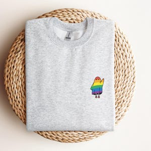May include: A pale grey sweatshirt featuring a small, embroidered ghost design. The ghost is wearing a rainbow cloak and has tiny feet. The sweatshirt is presented on a woven, circular mat.