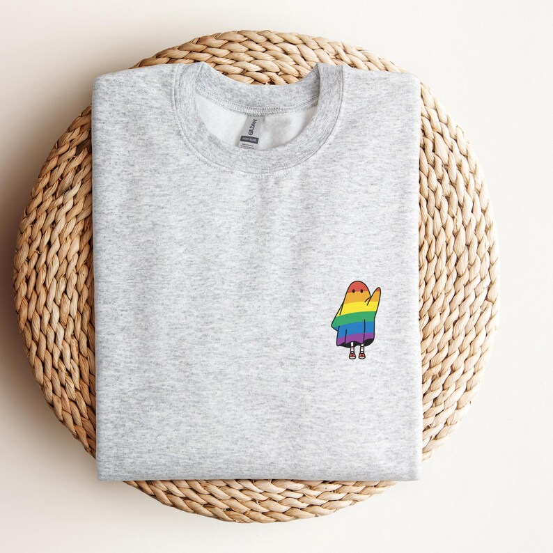 May include: A pale grey sweatshirt featuring a small, embroidered ghost design. The ghost is wearing a rainbow cloak and has tiny feet. The sweatshirt is presented on a woven, circular mat.