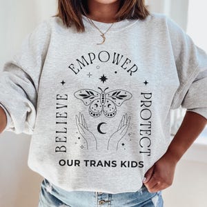 Può includere: Felpa grigio chiaro con le parole nere "EMPOWER BELIEVE PROTECT OUR TRANS KIDS". Al centro c'è una grafica di farfalla e mani. La felpa è in stile casual e confortevole.