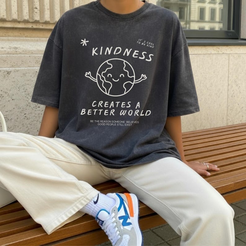 May include: Charcoal grey t-shirt featuring the word "KINDNESS" and a smiling globe graphic. The text "CREATES A BETTER WORLD" is below. The shirt is worn with cream trousers and white trainers with blue and orange details.