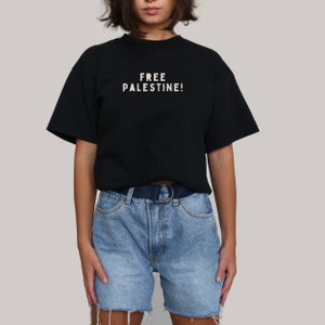 K&ouml;nnte beinhalten: Ein schwarzes T-Shirt mit dem Aufdruck "FREE PALESTINE!" in wei&szlig; auf der Vorderseite. Das Shirt wird von einer Person mit kurzen braunen Haaren getragen und zu einer hellblauen Jeans-Shorts kombiniert.