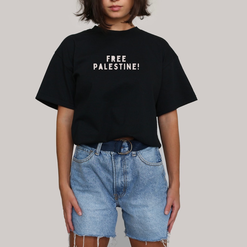 K&ouml;nnte beinhalten: Ein schwarzes T-Shirt mit dem Aufdruck "FREE PALESTINE!" in wei&szlig; auf der Vorderseite. Das Shirt wird von einer Person mit kurzen braunen Haaren getragen und zu einer hellblauen Jeans-Shorts kombiniert.