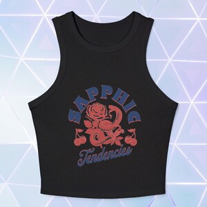 Puede incluir: Top corto negro con un dise&ntilde;o gr&aacute;fico. El dise&ntilde;o presenta la palabra "Sapphic" en azul, arqueada sobre una ilustraci&oacute;n de rosa y serpiente roja, con la palabra "Tendencies" debajo. Las cerezas est&aacute;n a ambos lados de la ilustraci&oacute;n.