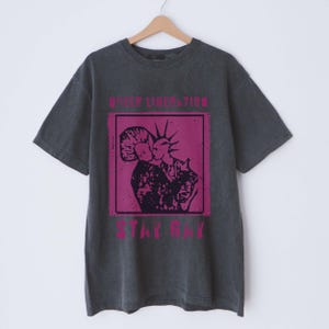 May include: A dark gray t-shirt with a pink graphic print of two people kissing. The text "Queer Liberation" and "Stay Gay" is printed in pink on the shirt.
