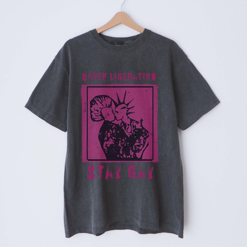 May include: A dark gray t-shirt with a pink graphic print of two people kissing. The text "Queer Liberation" and "Stay Gay" is printed in pink on the shirt.