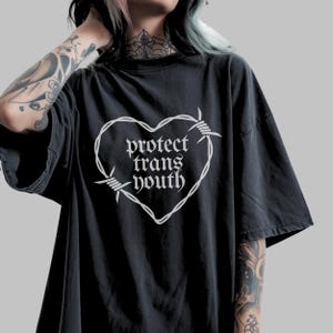 以下が含まれることがあります： 黒いTシャツ。白いハート型のデザインは有刺鉄線で作られています。「protect trans youth」の文字がゴシック体でハートの中にプリントされています。Tシャツは半袖です。