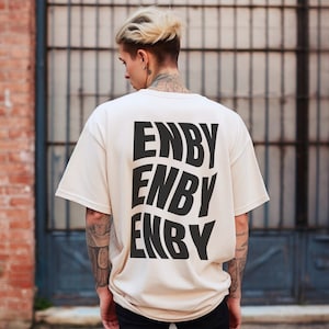 Könnte beinhalten: Ein beiges T-Shirt mit dem Wort "ENBY", das dreimal in einer schwarzen, fetten, stilisierten Schrift wiederholt wird.