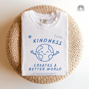 May include: White t-shirt featuring a blue illustration of a smiling globe with arms, the word "KINDNESS", and the phrase "CREATES A BETTER WORLD". The shirt also has the text "IT'S COOL TO BE KIND" and "BE THE REASON SOMEONE BELIEVES GOOD PEOPLE STILL EXIST".