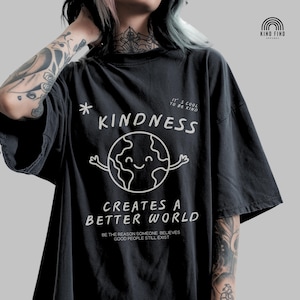 May include: Dark grey t-shirt featuring a white graphic of a smiling Earth and the words "KINDNESS CREATES A BETTER WORLD". Additional text includes "IT'S COOL TO BE KIND" and "BE THE REASON SOMEONE BELIEVES GOOD PEOPLE STILL EXIST".