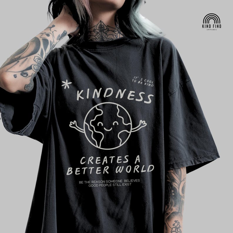 May include: Dark grey t-shirt featuring a white graphic of a smiling Earth and the words "KINDNESS CREATES A BETTER WORLD". Additional text includes "IT'S COOL TO BE KIND" and "BE THE REASON SOMEONE BELIEVES GOOD PEOPLE STILL EXIST".
