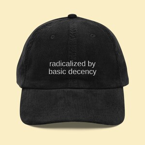 Puede incluir: Gorra de b&eacute;isbol de pana negra con la frase blanca "radicalized by basic decency". La gorra tiene una visera curva y un bot&oacute;n en la parte superior.