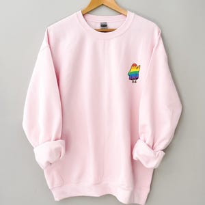May include: Pale pink sweatshirt with a crew neck and long sleeves. A small, colourful cartoon character in rainbow colours is embroidered on the left chest. The sweatshirt is hanging on a wooden hanger.