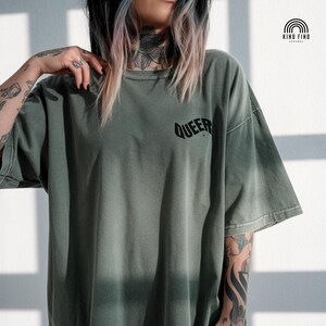 May include: Olive green t-shirt with the word "QUEER" printed in black on the upper right chest. The shirt has short sleeves and a relaxed fit. The Kind Find Apparel logo is in the upper right corner.