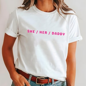 Puede incluir: Camiseta blanca con mangas enrolladas. La camiseta presenta el texto "SHE / HER / DADDY" en rosa brillante, centrado en la parte delantera. El modelo lleva vaqueros azules y un cinturón marrón.