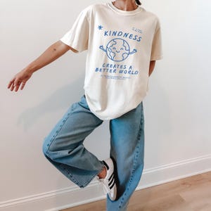 May include: Cream t-shirt featuring the phrase "Kindness Creates a Better World" in blue, with a smiling globe graphic. The person is wearing light blue jeans and white trainers with black stripes.