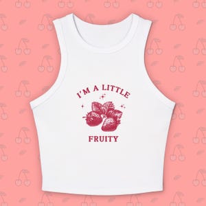 Op de afbeelding: Witte crop top met een ronde hals en een rode afbeelding van aardbeien en de tekst "I'M A LITTLE FRUITY". De top is mouwloos en gemaakt van een zacht materiaal. De achtergrond is roze met een kersenpatroon.