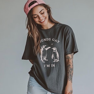 May include: A dark gray t-shirt with a graphic of two penguins dancing and the text "Sounds Gay I'm In". The model is wearing a pink baseball cap and blue jeans.