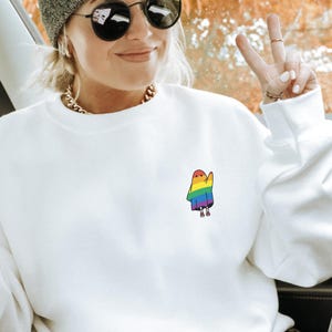 May include: White sweatshirt featuring a small, rainbow-coloured ghost graphic on the chest. The sweatshirt has a crew neck and long sleeves. The ghost design is in the colours of the rainbow.