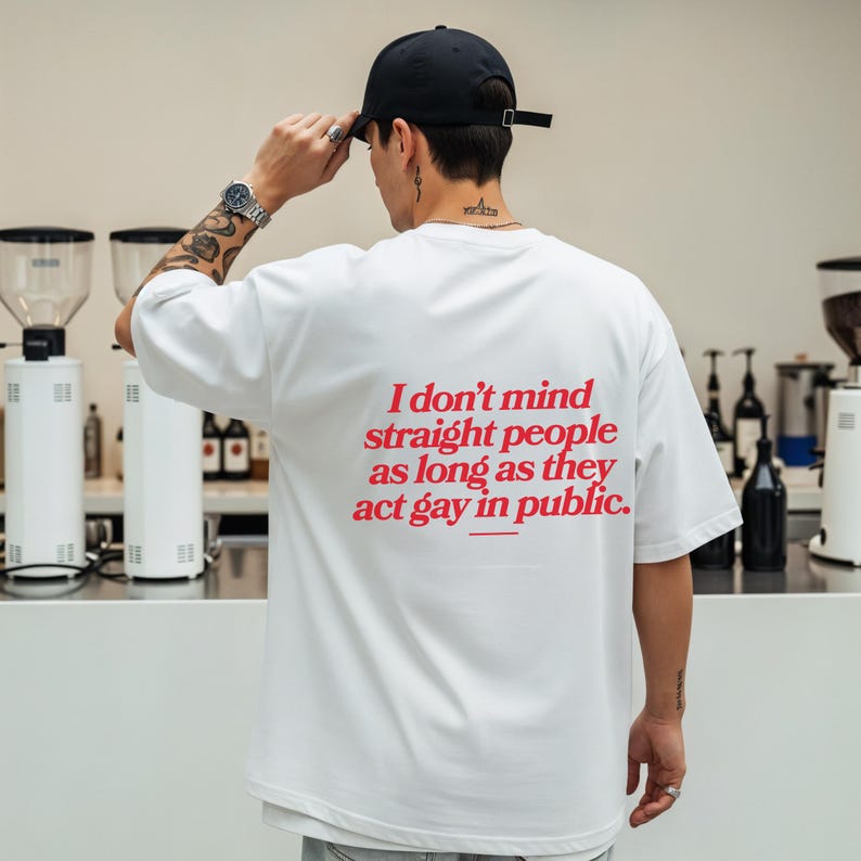 May include: White t-shirt with the text "I don't mind straight people as long as they act gay in public" in red. The shirt is worn with a black baseball cap and a silver watch.