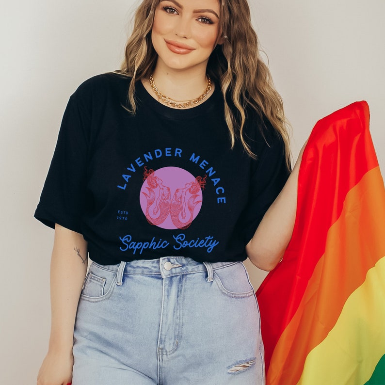 May include: A person wearing a black t-shirt with a purple and blue graphic of two mermaids. The graphic says "Lavender Menace" and "Sapphic Society". The person is holding a rainbow pride flag.