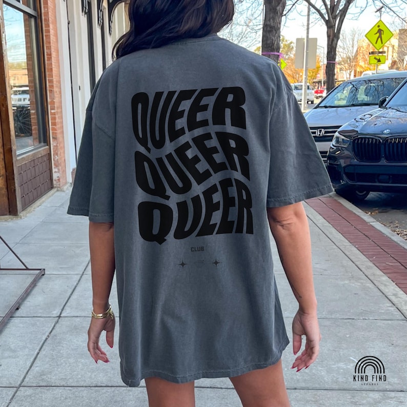 May include: Gray t-shirt with the word "QUEER" stacked four times in a wavy, black font. The word "CLUB" and two small star-like symbols are below the word. The shirt is worn by a person on a city sidewalk.
