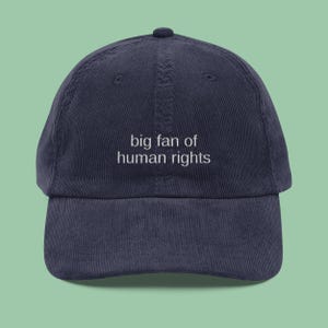 May include: A navy blue corduroy baseball cap featuring the embroidered phrase "big fan of human rights" in white. The cap has a curved peak and a fabric-covered button. A stylish, casual accessory.