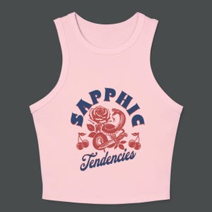 Puede incluir: Top corto rosa claro con un dise&ntilde;o gr&aacute;fico. El dise&ntilde;o presenta la palabra "Sapphic" en texto azul marino arqueado sobre una rosa roja, una serpiente y cerezas. Las palabras "Tendencies" est&aacute;n debajo.