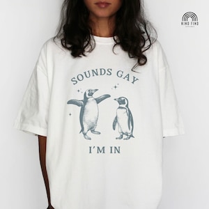 May include: White t-shirt with a graphic of two penguins and the text "Sounds Gay I'm In"