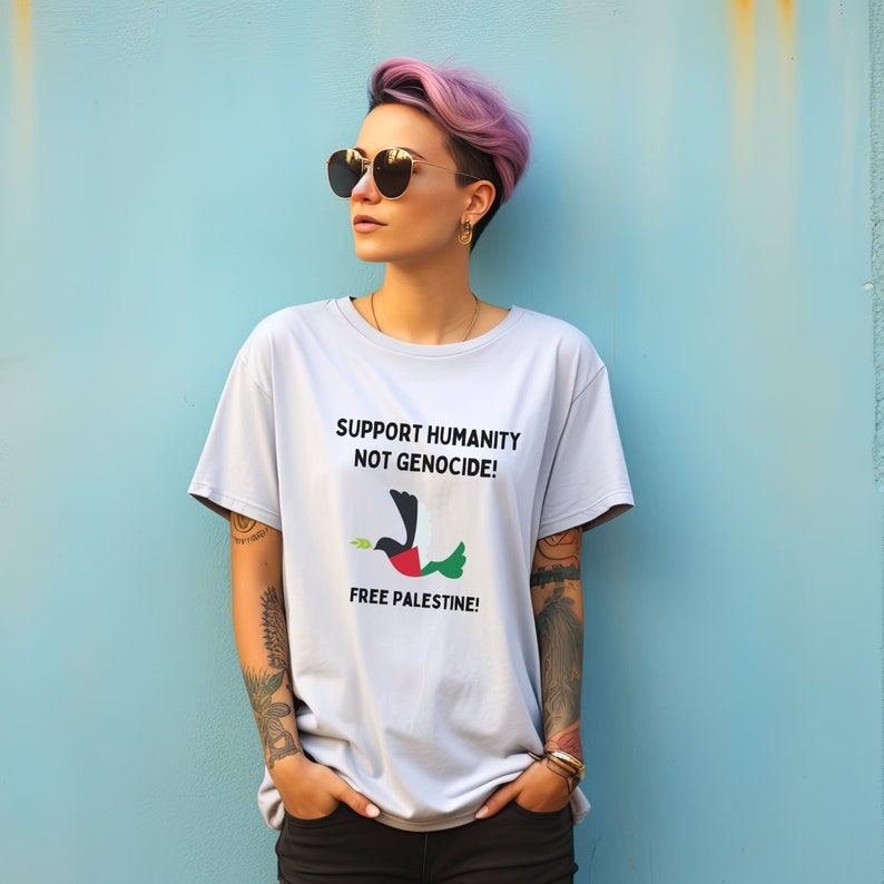 May include: A person wearing a light gray t-shirt with the text "Support Humanity Not Genocide!" and a black and green bird with a white olive branch in its beak. The text "Free Palestine!" is printed below the bird. The person has pink hair and is wearing sunglasses.