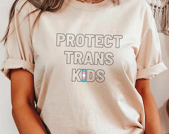 Può includere: Una maglietta beige con il testo "PROTECT TRANS KIDS" in lettere nere bordate. La parola "KIDS" ha un simbolo di bandiera transgender pride al centro delle lettere.