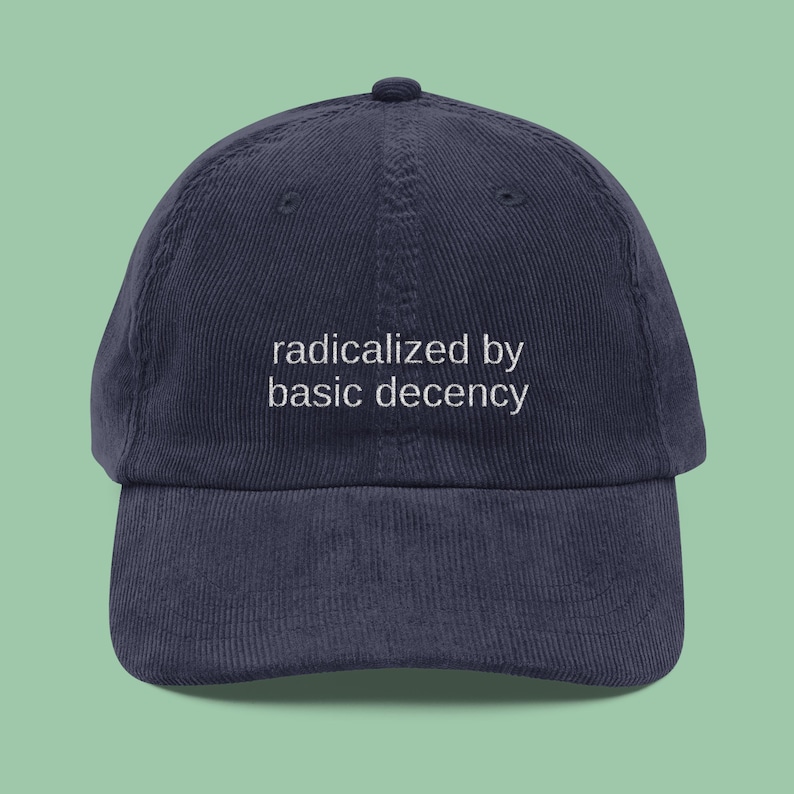 Puede incluir: Gorra de b&eacute;isbol de pana azul marino con la frase blanca "radicalized by basic decency". La gorra tiene una visera curva y un bot&oacute;n en la parte superior. El fondo es de color verde claro.