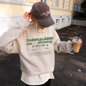 Op de afbeelding: Een beige sweatshirt met de tekst "RADICALIZED decency" in het groen, gecombineerd met een bruine baseballcap met "La Paris" in het wit. De sweatshirt heeft ook extra tekst en afbeeldingen. Een drankje in een doorzichtige beker wordt in de hand gehouden.